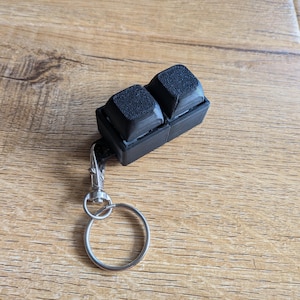 May include: A black mechanical keyboard switch keyring featuring two square, textured keycaps mounted on a rectangular base. The device includes a silver-coloured metal lobster clasp and a circular split ring for attachment to keys or bags.