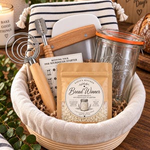 May include: A sourdough bread making kit in a round woven basket containing a wire dough whisk with a wooden handle, a metal lame, a glass jar with an orange seal, and a pouch labeled Kacie's Kitchen Bread Winner Dehydrated Sourdough Starter.