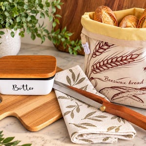 May include: A white ceramic butter dish with a wooden lid, a wooden cutting board, a bread knife with a wooden handle, a folded cloth napkin with a leaf pattern, and a beige fabric beeswax bread bag with wheat and bee illustrations.