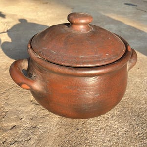 May include: A round, dark brown clay pot with a matching lid and two side handles. The surface of the vessel has a natural, earthy texture with visible variations in color and tone, featuring a simple knob handle centered on the lid.