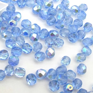 May include: A scattering of light blue, faceted beads with iridescent highlights. The beads are scattered across a white surface, with some beads partially contained in a small, white container. These beads are suitable for jewelry making and crafts.