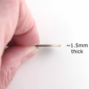 以下が含まれることがあります： 約1.5mmの厚さの薄い金属製の物体が指の間に挟まれています。その物体は硬貨または同様の円盤状のアイテムのようです。「~1.5mm thick」というテキストが物体の横に表示されています。