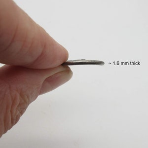 May include: A silver-colored metal disc, approximately 1.6 mm thick. The disc is being held by a finger.