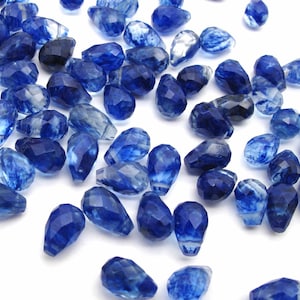 May include: A collection of blue faceted teardrop-shaped glass beads. The beads have a translucent, cloudy appearance with streaks of white.