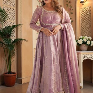 May include: A floor-length lavender anarkali dress featuring long sleeves, a square neckline, and intricate silver embroidery along the bodice and hem. The garment includes a matching sheer lavender dupatta draped over one shoulder with silver detailing.