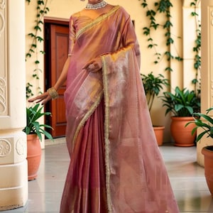 May include: A mauve-coloured saree featuring a sheer, lightweight fabric with a subtle sheen. The garment is adorned with a wide, intricate gold metallic border along the edges and a matching gold-toned blouse with short sleeves.
