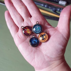 Könnte beinhalten: Vier runde Glasanhänger mit bronzenen Metallrahmen, jeder mit einem anderen Wappen des Hogwarts-Hauses: Gryffindor, Slytherin, Hufflepuff und Ravenclaw.