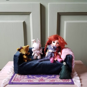 Op de afbeelding: Een miniatuurscène met een marineblauwe fluwelen bank met een teddybeer, een konijn, een pop en een vilten kat. Een kleine groene vilten kerstboom en een roze tapijt maken de setting compleet.