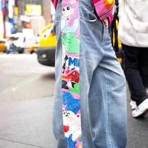 May include: Light blue denim jeans featuring a wide vertical panel on the side decorated with colorful anime-style illustrations, including a character with pink hair, the text "MILK," a red bird, and a white cat-like figure with blue accents.