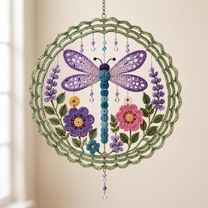 Op de afbeelding: Een ronde gehaakte wandhanger met een centrale libel met paarse en blauwe vleugels en een blauw lichaam. Het ontwerp bevat gehaakte paarse, roze en gele bloemen met groene bladeren, omlijst door een geschulpte groene rand met hangende kristallen kralen.