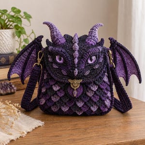 Puede incluir: Un bolso bandolera de ganchillo con forma de cabeza de dragón, con escamas de lana morada y azul oscuro, dos cuernos y dos alas. El bolso cuenta con dos ojos morados, un cierre metálico de color bronce y una correa a juego con clips.