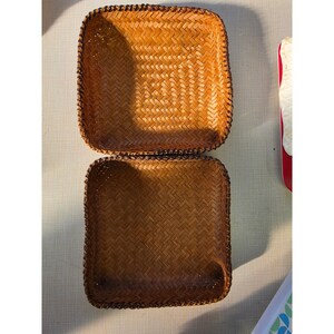 May include: Two square, shallow baskets crafted from woven brown natural fibers. Each basket features a consistent herringbone weave pattern throughout the base and sides, with a reinforced, darker braided rim along the top edges.
