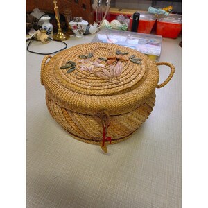 May include: A round, woven straw basket with a lid featuring a decorative floral appliqué in shades of tan, brown, and green. The basket has two side handles and a small, dangling red string tag attached to the front closure mechanism.