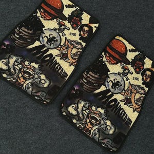 May include: Two rectangular car floor mats featuring a collage of anime-style illustrations, including a character in a straw hat, a laughing sun, and a figure surrounded by clouds. The text MONKEY D. LUFFY and D.VOID is printed on the mats.