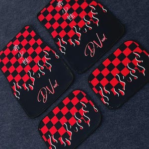 Puede incluir: Un juego de cuatro alfombrillas para coche con un patrón de cuadros rojos y negros, gráficos de llamas blancas y el texto "D.Void" en letra roja. Dos alfombrillas incluyen caracteres japoneses verticales en blanco, las otras dos solo llamas.