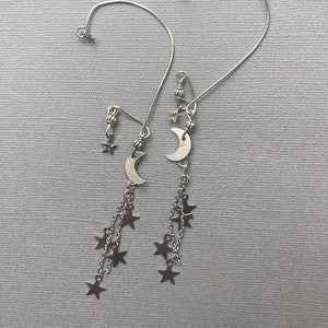 Puede incluir: Un par de pendientes de plata con un dije de luna creciente y dijes de estrellas colgantes. Los pendientes están hechos de alambre delgado y tienen un diseño simple.