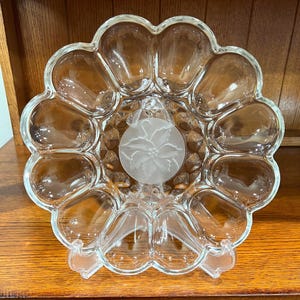 May include: A clear glass egg serving platter featuring twelve rounded indentations arranged in a circular pattern. The center of the platter displays a frosted circular medallion with an etched poinsettia flower design and scalloped edges.