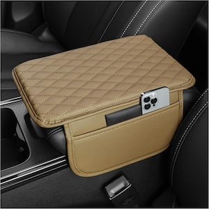 May include: A tan car center console armrest cover featuring a diamond-quilted pattern on the top surface. The cover includes an integrated side pocket containing a silver smartphone. The accessory is designed to fit over a vehicle armrest.