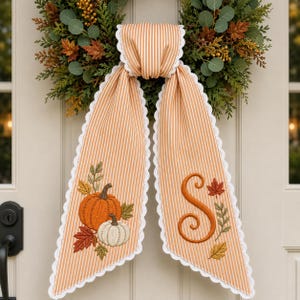 May include: A fabric door hanger featuring orange and white vertical stripes with a white scalloped edge. The left side displays embroidered pumpkins and autumn leaves, while the right side features a large embroidered letter S and fall foliage.