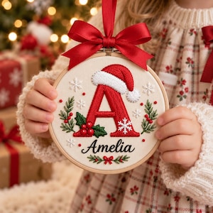 May include: A circular wooden embroidery hoop featuring a cream-colored fabric background. The design includes a large red embroidered letter A topped with a Santa hat, surrounded by green holly leaves, red berries, and white snowflakes. The name Amelia is stitched in black script below, finished with a red ribbon bow.
