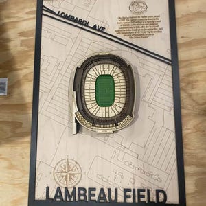 May include: A rectangular wooden wall art piece featuring a layered 3D model of Lambeau Field with a green football field. The background shows a map with the text Lombardi Ave and Lambeau Field, including a compass rose and historical text.