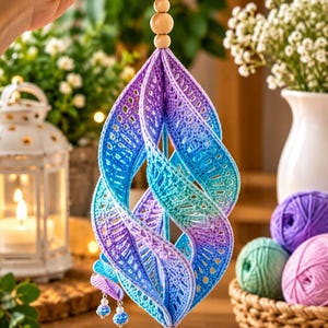 Könnte beinhalten: Ein spiralförmiges Häkelornament mit einem Farbverlauf aus violettem, blauem und türkisfarbenem Garn. Das Stück hängt an einer weißen Schnur mit zwei hellen Holzperlen oben und unten und ist mit einer kleinen gehäkelten Kugel und silbernen Akzenten versehen.
