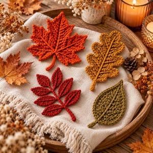 Puede incluir: Cuatro hojas de ganchillo en colores otoñales: una hoja de arce naranja grande, una hoja de roble amarillo dorado, una hoja compuesta de color rojo oscuro con varios folíolos y una hoja verde oliva, sobre un paño de tela beige.