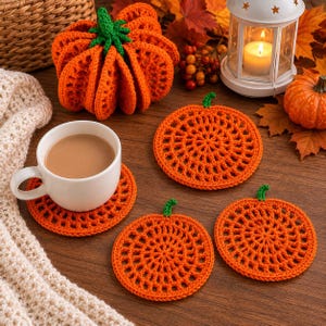 May include: A set of three round orange crochet pumpkin coasters with green stems, a larger three-dimensional crochet pumpkin, a white ceramic mug filled with coffee, and a white lantern on a wooden surface with autumn leaves and a knit blanket.