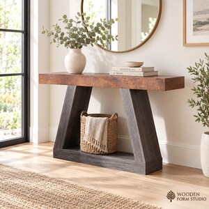 May include: A console table featuring a rectangular brown wood-grain top supported by two dark gray angled legs connected by a base. A woven basket with a light-colored cloth sits on the base, while a vase with greenery and books rest on top.