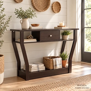 May include: A dark brown wooden console table featuring a single central drawer with a silver knob, an open storage shelf above, and a bottom shelf. The table has angled legs and is styled with books, a ceramic vase, a woven basket, and plants.