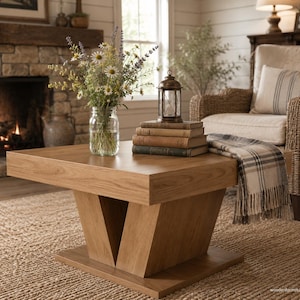 May include: A square wooden coffee table with a thick top and a V-shaped pedestal base. On the table sits a clear glass jar filled with wildflowers, a small metal lantern, and a stack of three vintage books with a plaid throw blanket draped over.