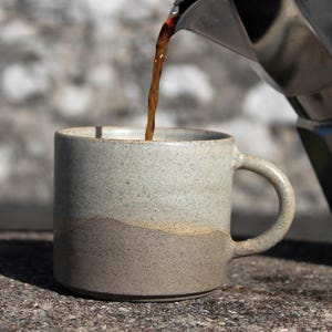 Op de afbeelding: Een tweekleurige keramische mok met een gespikkeld lichtgrijs bovengedeelte en een donkerbruine onderkant. Een metalen Bialetti mokkapot schenkt donkere koffie in de mok. De mok heeft een eenvoudige cilindrische vorm met een gebogen handvat.