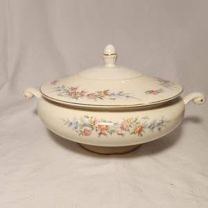 May include: A cream-colored ceramic tureen with a matching lid, featuring a delicate floral pattern of pink, yellow, and blue flowers with green leaves. The tureen has two curved side handles and a gold-trimmed rim and lid finial.