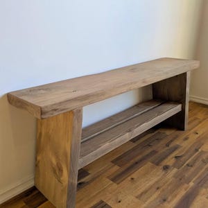 Può includere: Una panca lunga e rettangolare in legno con una superficie superiore spessa e piatta e un ripiano inferiore, sostenuta da due pannelli laterali solidi e inclinati. La panca è realizzata in legno marrone chiaro con venature e nodi visibili.