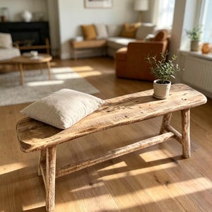 Puede incluir: Un banco rectangular de madera con un acabado natural desgastado y cuatro patas robustas conectadas por un travesaño. Un cojín rectangular de tela beige claro descansa en el lado izquierdo y una pequeña planta verde en una maceta está a la derecha.