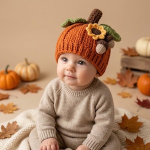 May include: A crochet pumpkin hat in vibrant orange with a ribbed texture, featuring a brown stem, two green leaves, a yellow sunflower, and two small acorns attached to the side. The hat is worn by a baby dressed in a beige long-sleeve sweater.