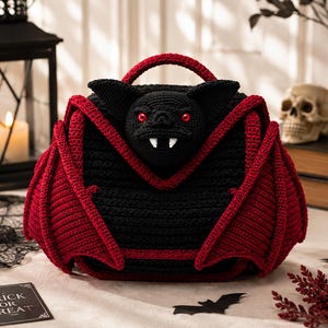 May include: A crochet handbag shaped like a bat with a black body, red wings, and a central bat head featuring red eyes and white fangs. The bag has a top handle and is constructed from yarn with a visible stitch texture throughout the design.