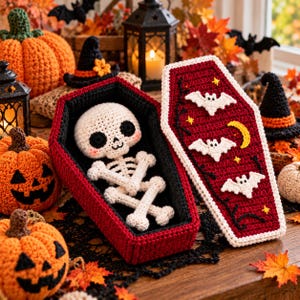May include: A crocheted white skeleton doll with black eyes and pink cheeks resting inside a red and black coffin. The coffin lid is decorated with three white crocheted bats, a yellow crescent moon, and small yellow stars on a red background.