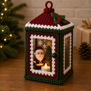May include: A decorative crochet lantern featuring a dark green and red frame with white trim. The top is adorned with a green holly leaf and red berries. Inside, a crocheted Santa Claus and a small Christmas tree stand beside a lit candle.