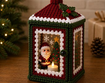 May include: A decorative crochet lantern featuring a dark green and red frame with white trim. The top is adorned with a green holly leaf and red berries. Inside, a crocheted Santa Claus and a small Christmas tree stand beside a lit candle.