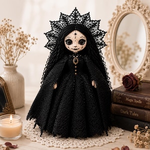 May include: A handcrafted crochet doll featuring a pale face with detailed black facial markings, large dark eyes, and long black hair. The doll wears a black lace-trimmed gown and a matching intricate black lace headpiece or halo.