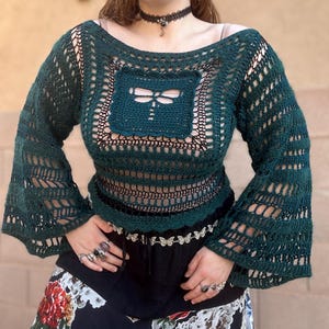 May include: A dark green crochet long-sleeve top featuring an open-knit pattern and a central square motif with a dragonfly design. The garment has a wide boat neckline and flared sleeves, layered over a black base garment.