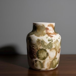 Może przedstawiać: Ceramiczny wazon z szeroką, krótką szyjką i zaokrąglonym korpusem. Powierzchnia posiada teksturowany, nakrapiany wzór nakładających się na siebie okrągłych kształtów w odcieniach oliwkowej zieleni, ziemistego brązu i kremu, tworząc zróżnicowany wygląd.