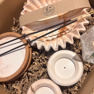 Könnte beinhalten: Ein Set aus Wohnaccessoires, bestehend aus einer großen Schale mit gewelltem Rand und einem pfirsichfarben-weißen Marmormuster, umwickelt mit einer Papierbanderole mit der Aufschrift Olive.waves. Zwei schwarze Räucherstäbchen und zwei kleine, runde Schalen.