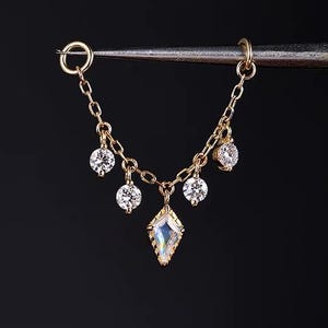 May include: A gold chain connector earring featuring four round clear stones and one central kite-shaped iridescent stone. The delicate gold links form a draped design, with each stone suspended from the chain, creating a symmetrical arrangement.