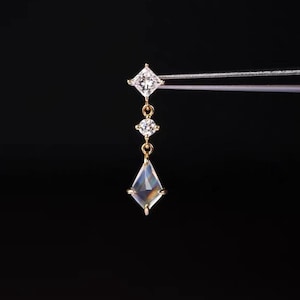 May include: A gold dangle earring featuring a princess-cut clear gemstone at the top, a small round brilliant-cut clear gemstone in the center, and a kite-shaped iridescent gemstone at the bottom, all set in a delicate gold metal chain structure.