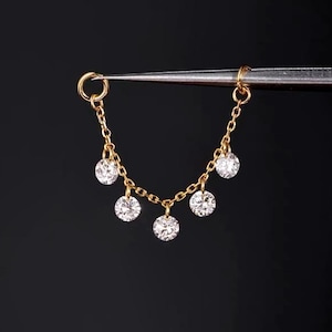 May include: A gold-toned jewelry connector chain featuring five small, round, clear faceted stones suspended from delicate links. The piece is held by metal tweezers, showcasing the symmetrical arrangement of the stones hanging in a curved line.