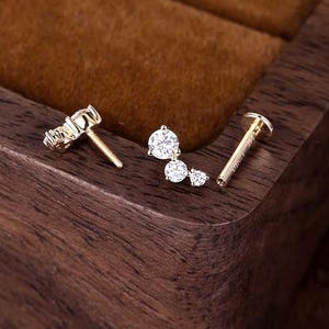 May include: A gold flat back stud earring featuring three round clear stones arranged in a cluster. The earring has a threaded post and a circular flat back piece. The stones are set in a gold metal frame, showcasing a bright, reflective finish.