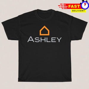May include: A black short-sleeved t-shirt featuring an orange house-shaped icon centered above the word ASHLEY printed in light gray capital letters. The shirt has a crew neckline and a standard fit, displaying a clean and simple graphic design.