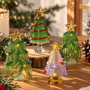 Puede incluir: Cuatro figuras de árboles de Navidad de cristal con estrellas doradas en la punta. Tres árboles son verdes con hebras de cristal entrelazadas y cuentas de colores. Un árbol es transparente con diseño en espiral y cuentas sobre base dorada.